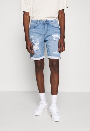 Pantalones cortos de mezclilla azul claro con detalles desgastados, puños enrollados, diseño de cinco bolsillos y cierre de botón, combinados con una camiseta beige y zapatillas blancas.