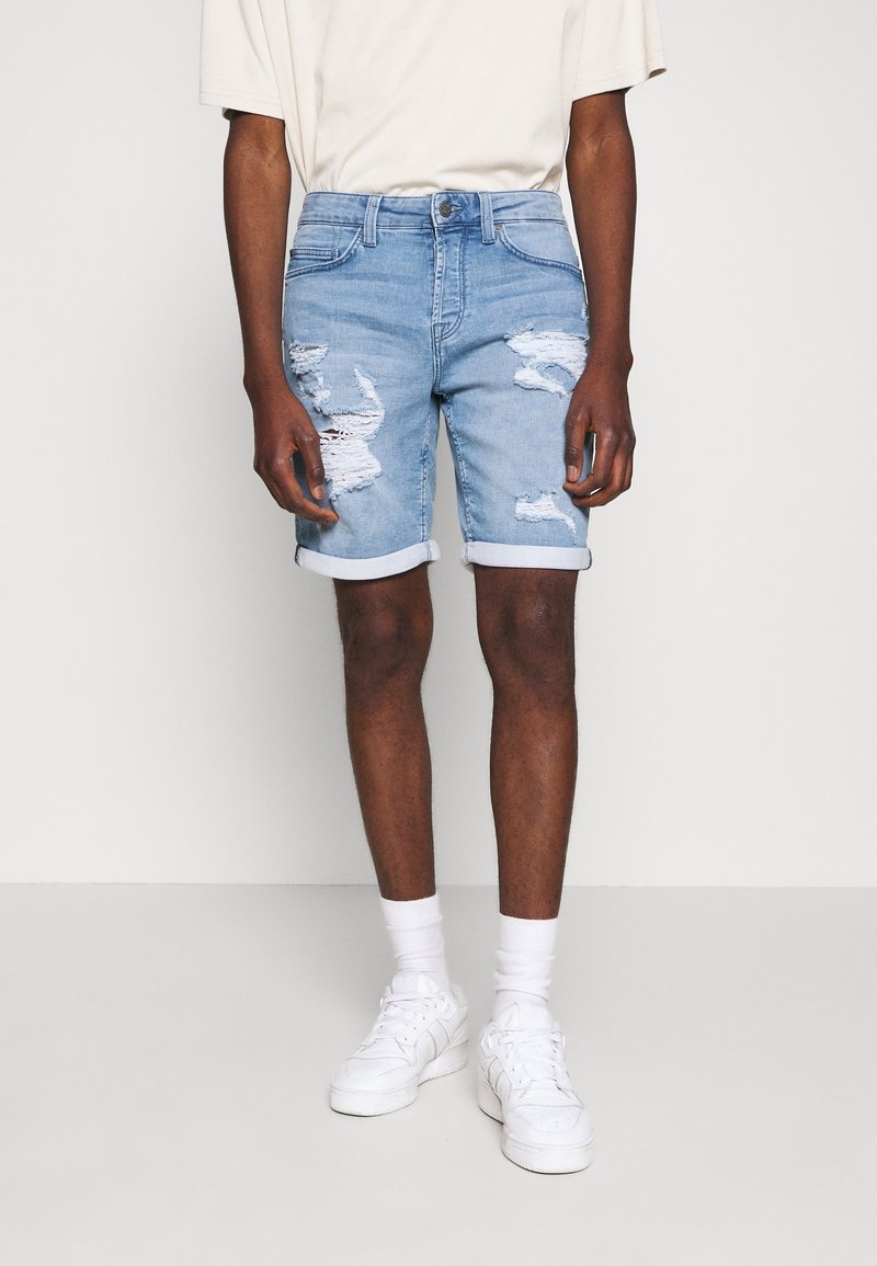 Pantalones cortos de mezclilla azul claro con detalles desgastados, puños enrollados, diseño de cinco bolsillos y cierre de botón, combinados con una camiseta beige y zapatillas blancas.