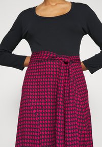 Svart långärmad topp som kombineras med en kjol i rosa och marinblå Hundtandsmönster, med knyt i midjan och en svävande design.