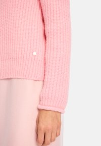 Rosa gestrickter Pullover mit gerippten Bündchen und Saum, der eine glatte Textur aufweist und einen kleinen dekorativen Knopf auf der unteren linken Seite hat.