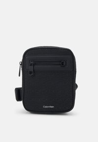 Zwarte schoudertas met een gestructureerd oppervlak, voorzien van een ritsvak aan de voorkant, een geweven logo-patroon en een verstelbare schouderband. Calvin Klein-merkzichtbaar.