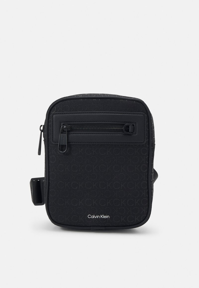 Zwarte schoudertas met een gestructureerd oppervlak, voorzien van een ritsvak aan de voorkant, een geweven logo-patroon en een verstelbare schouderband. Calvin Klein-merkzichtbaar.