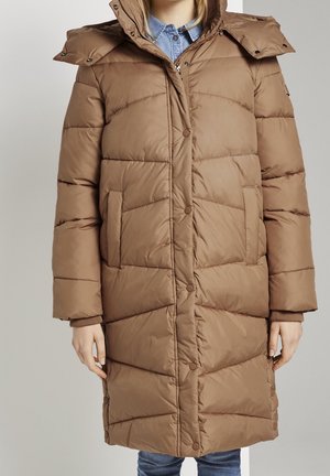 Frau trägt eine lange braune Steppjacke mit vorderen Druckknöpfen, einem Reißverschluss, seitlichen Taschen und abnehmbarer Kapuze über einem blauen Jeanshemd.