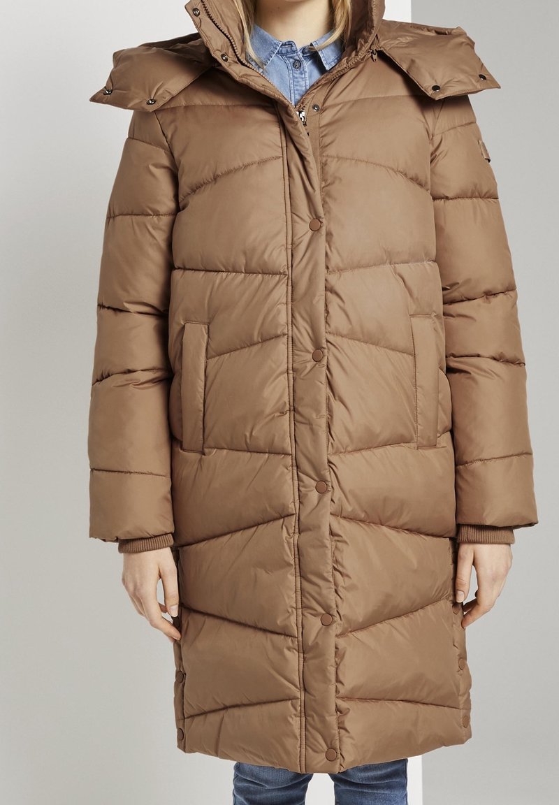 Femme portant un long manteau matelassé marron avec des boutons-pression à l'avant, une fermeture éclair, des poches latérales et une capuche amovible, sur une chemise en denim bleu.
