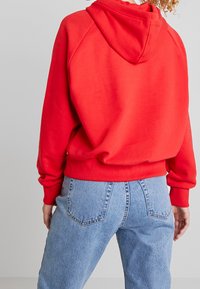 Persona con una sudadera roja brillante y jeans de tiro alto azul claro, vista desde atrás contra un fondo gris claro liso.