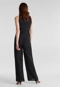 Femme aux longs cheveux bruns portant une combinaison noire sans manches et des sandales noires à talons, se tenant de dos sur un fond gris uni.