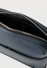 Δερμάτινη θήκη ναυτικού μπλε με ασημένιο φερμουάρ. Υφή επιφάνειας και μαύρη επένδυση ορατή στο εσωτερικό. Γωνιώδες σχήμα με λεπτό προφίλ.