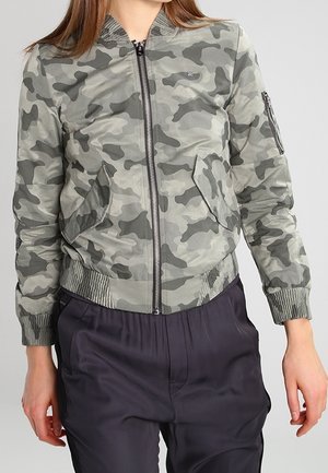 Frau trägt eine graue und grüne Camouflage-Bomberjacke mit Fronttaschen und schwarze Hose vor einem schlichten Hintergrund.