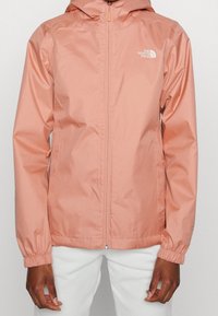 Chaqueta impermeable ligera de color melocotón con capucha, puños elásticos, cierre de cremallera y un pequeño logo en el pecho izquierdo. Textura lisa.