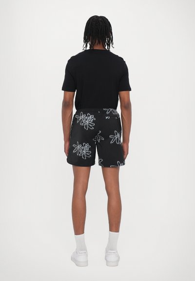 Schwarzes T-Shirt kombiniert mit gemusterten schwarzen Shorts mit weißer floralem Stickerei. Model trägt weiße Sneakers und Socken.