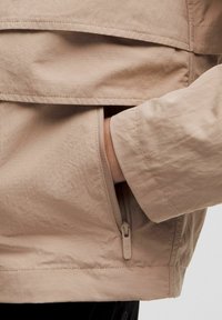 Main dans une poche zippée d'une veste légère beige, avec une couture et une texture de tissu visibles.