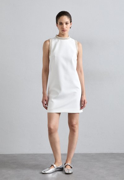 Weißes ärmelloses Kleid mit einer taillierten Silhouette und kunstvoll geflochtenem Ausschnitt. Kombiniert mit flachen Schuhen in metallic Silber.
