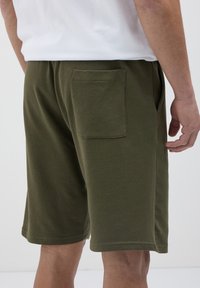 Homme portant un short olive vert mi-long avec une poche arrière et des poches latérales, associé à une chemise blanche.