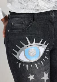 Jeans de mezclilla negra con un gran gráfico de ojo plateado y azul con rayos y estrellas en el muslo izquierdo. Incluye un diseño clásico de cinco bolsillos.