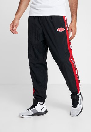 Mężczyzna noszący czarne spodnie dresowe z czerwono-białymi paskami po bokach oraz białe sportowe buty marki Nike, stoi na jednolitym tle.