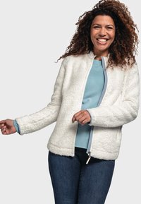 Veste en polaire blanche duveteuse avec une fermeture éclair à l'avant, un col rond et des manches longues. Superposée sur une chemise bleu clair et associée à un jean foncé.