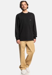 Černé triko s dlouhým rukávem a kapsou na hrudi, spárované s béžovými volnými kalhotami. Model má na sobě bílé boty; jednoduchý, neformální design.