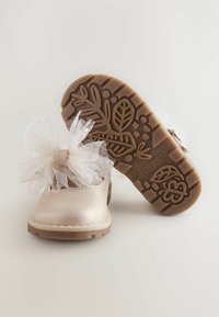 Helle rosa metallische Schuhe mit einer strukturierten Oberfläche, die eine große weiße Tüllschleife tragen. Die braune Gummisohle ist mit Blumenmustern versehen.