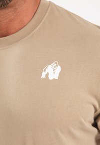 Šviesiai ruda trumpomis rankovėmis marškinėliai, pagaminti iš medvilnės, su maža balta gorilos grafika kairėje krūtinės pusėje.