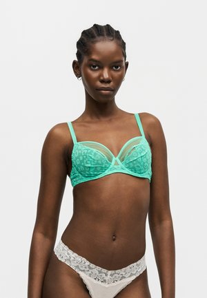 Femme aux cheveux tressés portant un soutien-gorge en dentelle vert menthe et une culotte en dentelle blanche, se tenant devant un fond clair uni.