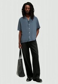 Uomo con dreadlocks che indossa una camicia blu a maniche corte, pantaloni neri a gamba larga, scarpe nere, mentre tiene una borsa a tracolla in pelle nera.