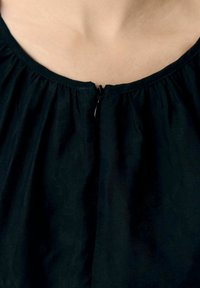Schwarze Bluse mit rundem Halsausschnitt, gerafftem Stoff und einem kleinen Reißverschluss vorne. Glatte Textur mit dezentem Muster.