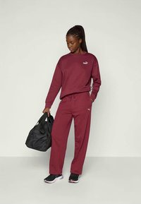 Burgunderfarget hettegenser og joggebukser laget av mykt stoff, med en liten hvit Puma-logo. Svart veske og joggesko fullfører antrekket.