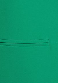 Zielony materiał o gładkiej teksturze, z poziomym detalem kieszeni. Materiał wydaje się elastyczny i dobrze dopasowany.