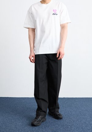 T-shirt en coton blanc avec un motif cœur rose et le texte "nike" sur le côté gauche de la poitrine, associée à un pantalon noir et des chaussures de sport noires.