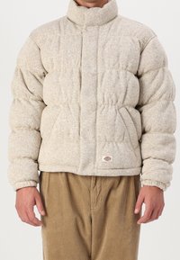 Puffjacka i beige, tillverkad av strukturerad tyg. Har en hög krage, elastiska mudd, och två sidofickor. Bärs över manchesterbyxor.
