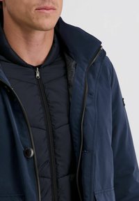 Man draagt gelaagde marineblauwe jassen met zichtbare ritsen en knopen, weergegeven van de nek tot het midden van de torso tegen een effen lichte achtergrond.