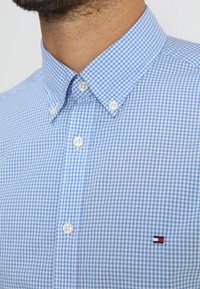 Camicia a righe blu e bianche con colletto appuntito, bottoni bianchi e una piccola etichetta con logo sul petto. Tessuto in cotone.