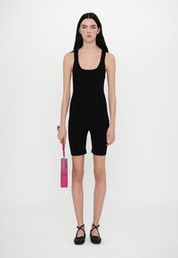 Bodysuit sans manches noir avec un design ajusté et une coupe short de vélo ; le modèle tient un sac à main rectangulaire rose avec une bandoulière en chaîne.