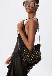 Borsa a tracolla in suede nero con borchie dorate e frange, dotata di una tracolla a catena. La modella indossa occhiali da sole oversize e un abito nero.