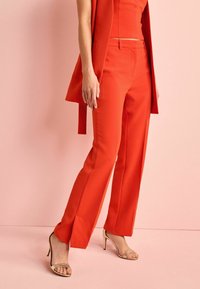 Red tailored pants with a straight cut, made from a smooth fabric, featuring side pockets and a low waistband. Paired with gold heels.