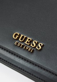 Zwarte leren portemonnee met een verhoogd gouden "GUESS" logo met daaronder "EST 1981". Gladde textuur met decoratieve stiksels langs de randen.