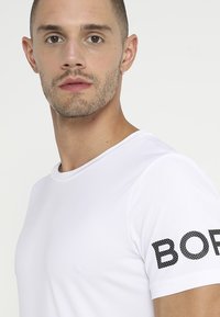 Camiseta blanca ajustada de manga corta. Presenta un escote redondeado y un texto impreso en negro con puntos "BORD" en la manga izquierda. Textura suave.