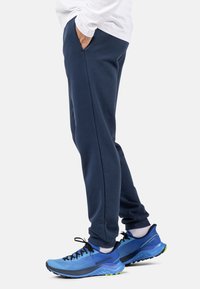 Marineblaue, schmal zulaufende Jogginghosen mit seitlichen Taschen, kombiniert mit blauen Sportschuhen, die schwarze Schnürsenkel und grüne Akzente haben.