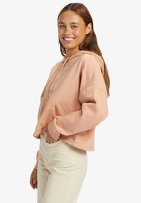 Sudadera cropped de suave tejido melocotón, con capucha ajustable, bolsillo frontal y puños acanalados. Combinada con pantalones de color claro.