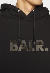 Sudadera negra con capucha ajustable; presenta un gran logotipo metálico "BALR." en un diseño elegante a rayas en el pecho.