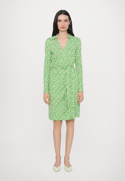 Robe verte à motifs de feuilles, à manches longues et avec une taille nouée. Le tissu a une texture lisse, associé à des chaussures de couleur claire.