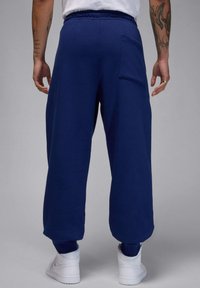 Person iført marineblå joggingbukser med baglomme, hvide sneakers, en hvid skjorte og synlige tatoveringer på begge arme.