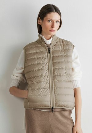 Kobieta o prostych brązowych włosach, ubrana w beżową pikowaną kamizelkę na białym dzianinowym swetrze i brązową spódnicę, stojąca na tle gładkiej jasnej ściany.