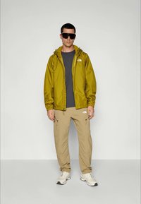 Homme portant des lunettes de soleil surdimensionnées noires, une veste olive, une chemise gris foncé, un pantalon cargo beige et des baskets blanches, se tenant contre un fond uni.
