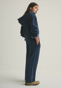 Felpa con cappuccio blu navy dalla vestibilità ampia, dettagli in denim su polsini e tasche, abbinata a pantaloni a gamba larga coordinati e scarpe beige.