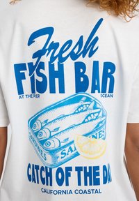White t-shirt featuring blue graphic print of a sardine can, text "Fresh Fish Bar" and "Catch of the Day," with a sliced lemon illustration.