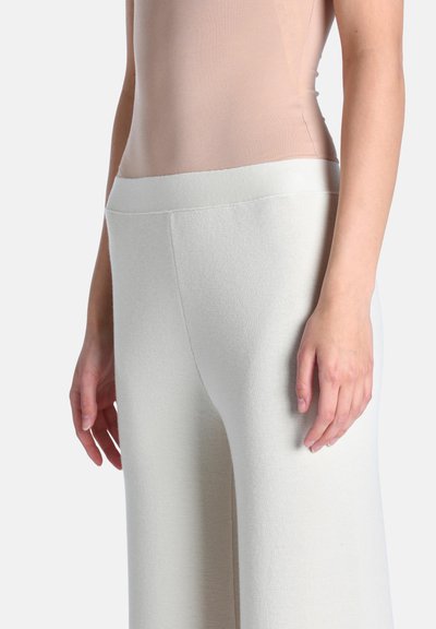 Personne portant un haut sans manches beige clair et un pantalon en maille blanc taille haute avec une large ceinture, mains détendues le long du corps.