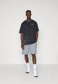Sort t-shirt med korte ærmer og farverigt sprøjtedesign i kanten, parret med grå shorts. Hvid Nike-logo på t-shirten. Hvide sneakers.
