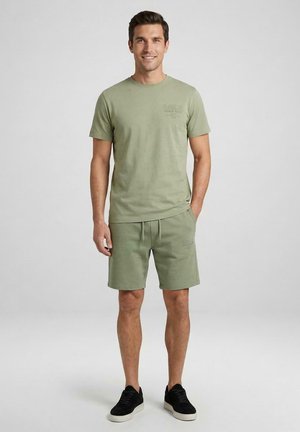 Man staat lachend, draagt een lichtgroen T-shirt met korte mouwen en bijpassende korte broek met zwarte sneakers tegen een effen achtergrond.