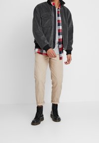 Chaqueta bomber de forro polar gris con cuello alto, ribete negro, dos bolsillos delanteros; lleva sobre una camisa de cuadros, camiseta gris y pantalones beige.